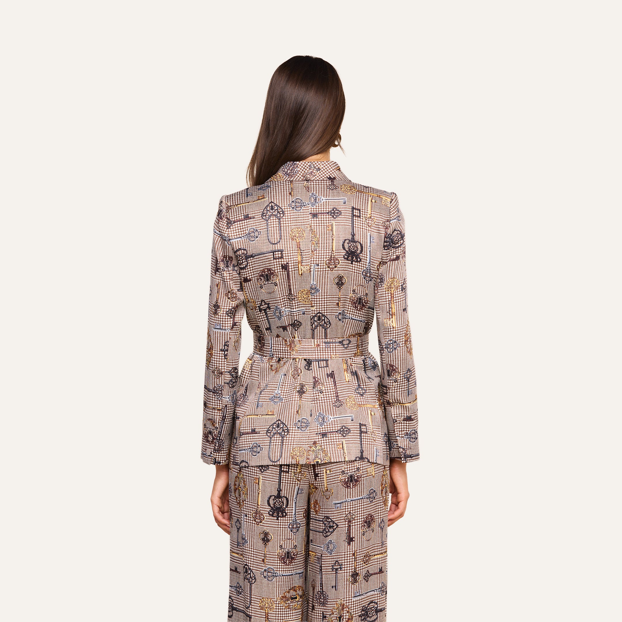 A woman stands facing away, wearing the Aven Wrap Blazer and matching wide-leg pants. The modern suiting set features a beige base with dark and light motifs, and her long brown hair is down.