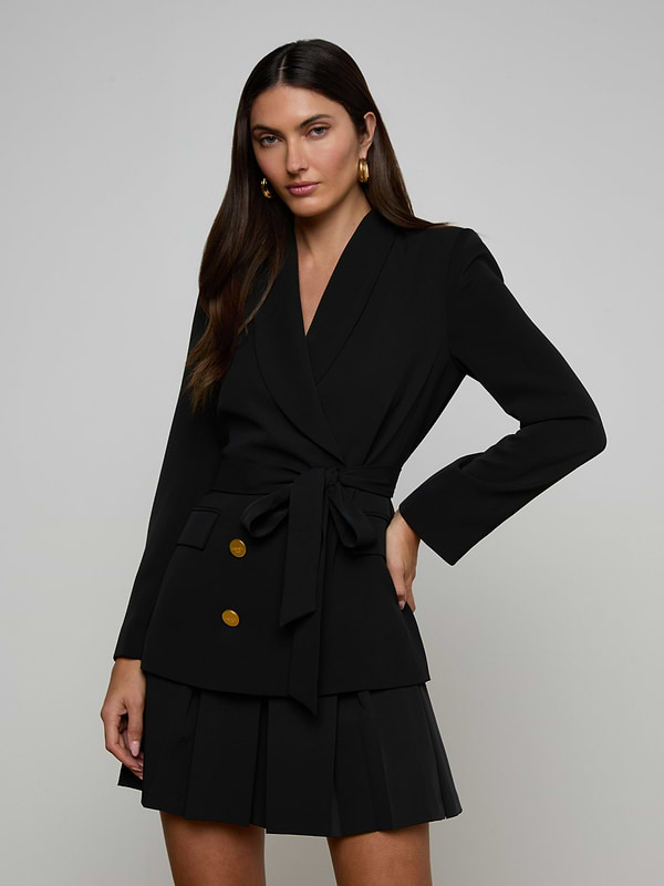 Woman wearing the Aven Wrap Blazer in black with gold buttons and a matching pleated skirt, standing with one hand on her hip against a plain light background—a modern suiting look.