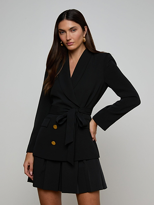 A woman with long dark hair wears the Aven Wrap Blazer—a black, gold-buttoned wrap-front blazer—paired with a pleated skirt. She stands against a plain light background, gazing confidently at the camera, one hand on her hip.
