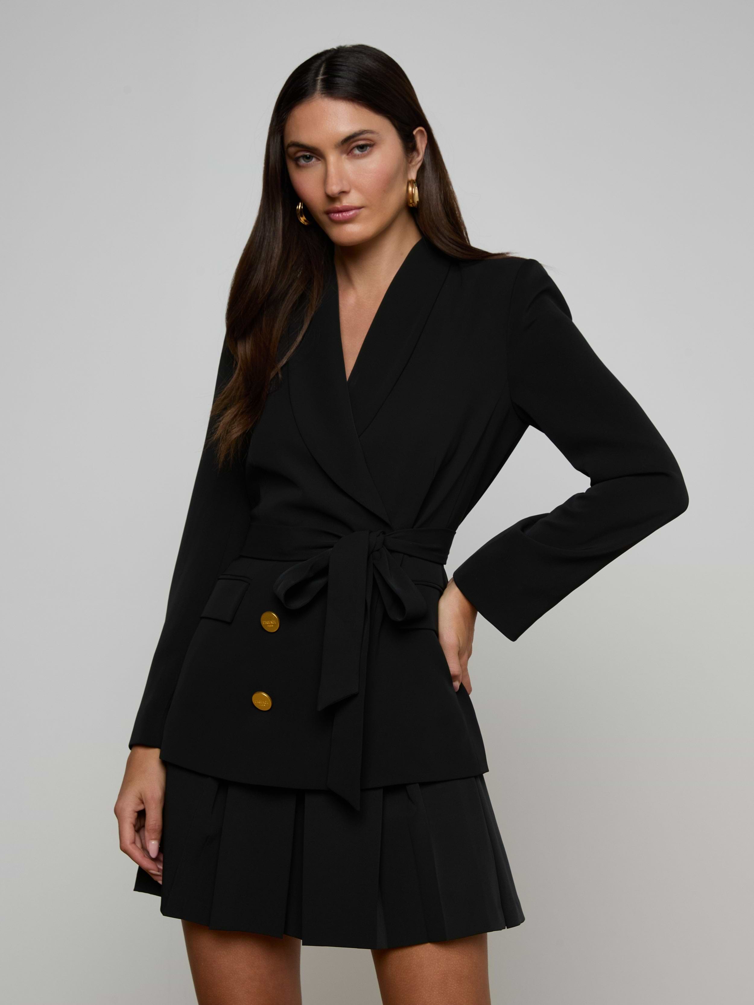 Woman wearing the Aven Wrap Blazer in black with gold buttons and a matching pleated skirt, standing with one hand on her hip against a plain light background—a modern suiting look.