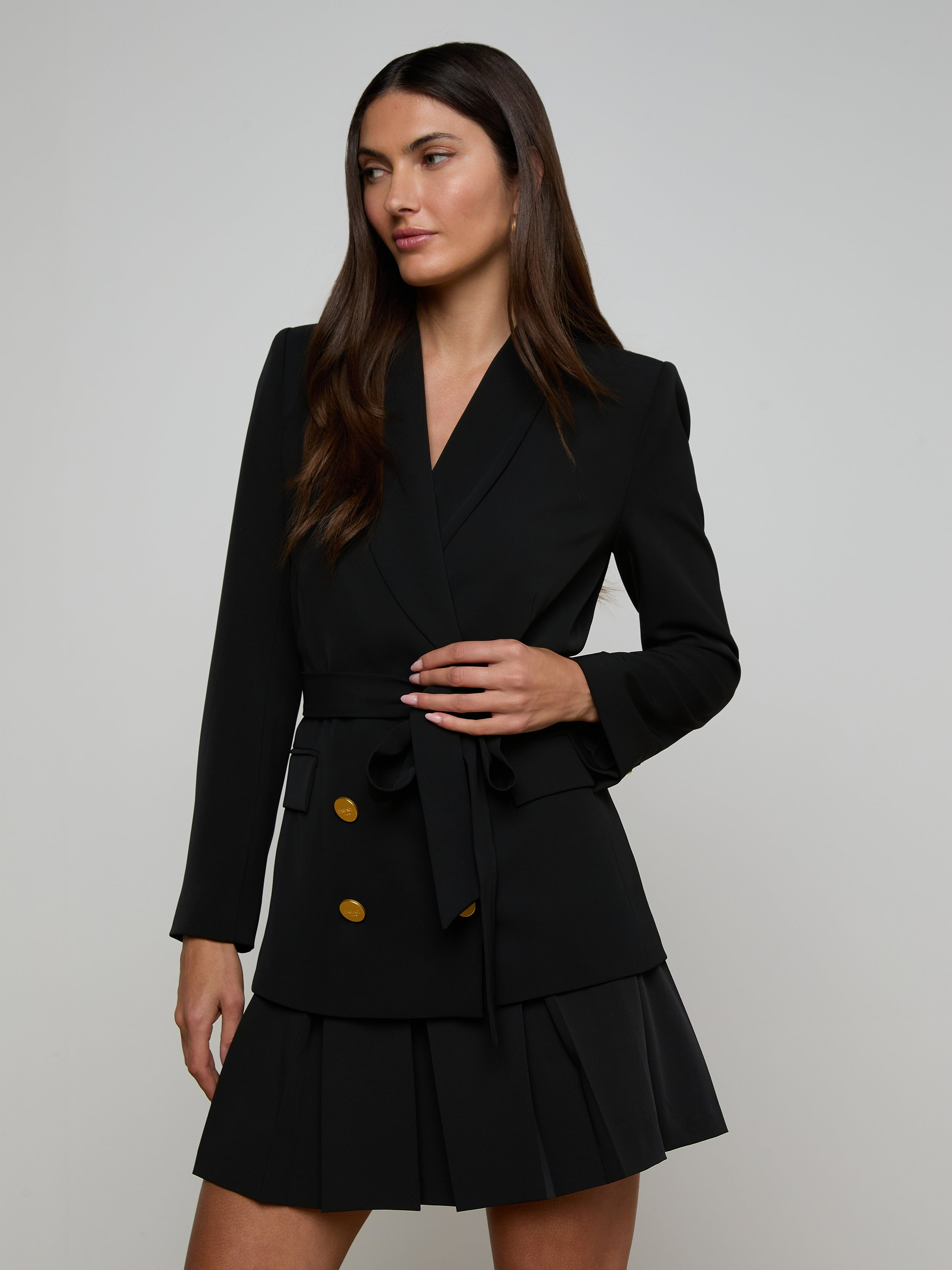 A woman wearing the Aven Wrap Blazer—a black wrap-front blazer dress with gold buttons and a pleated skirt—stands against a plain light background, looking to the side with one hand holding her waist belt.
