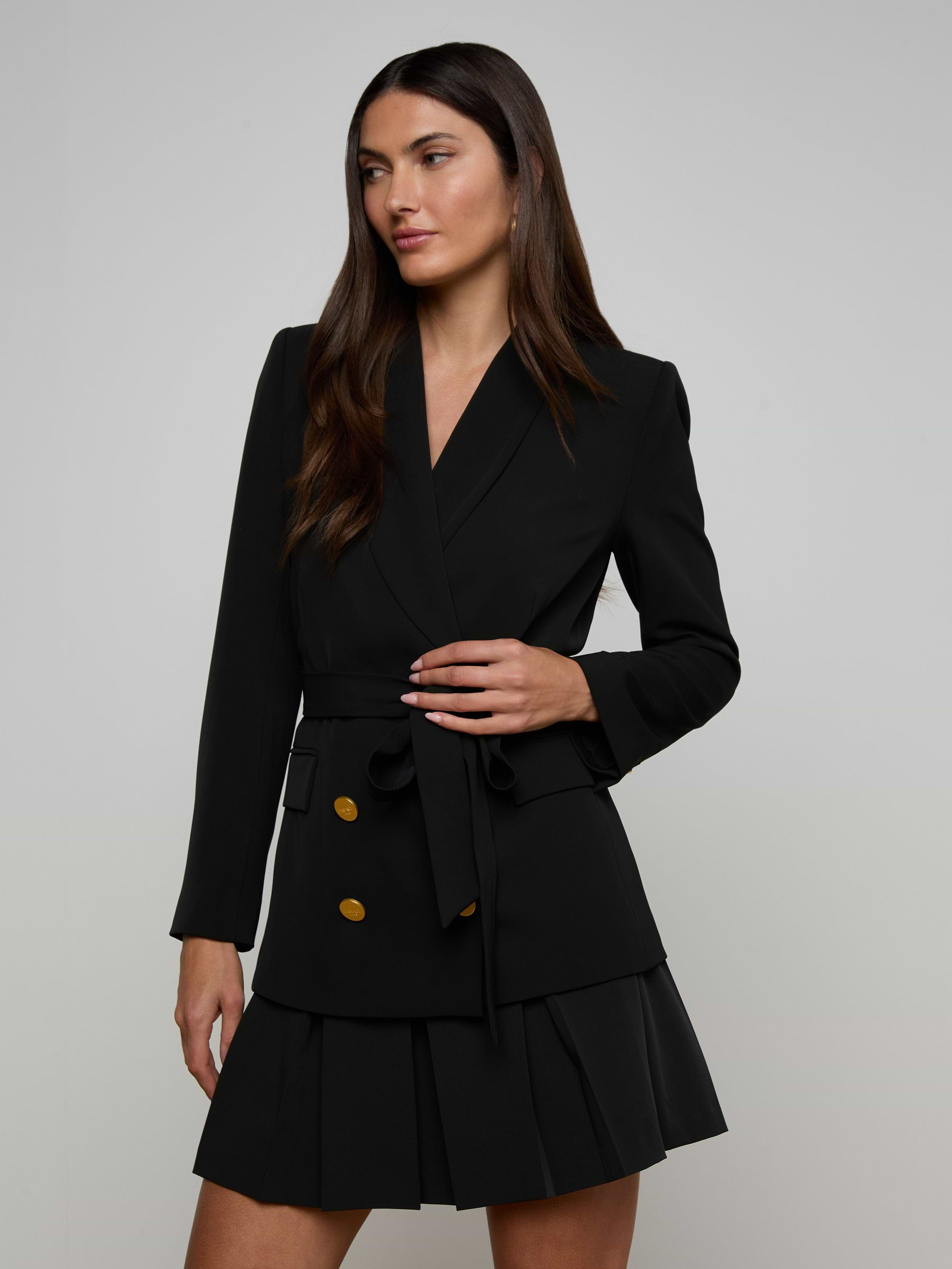 A woman wearing the Aven Wrap Blazer—a black wrap-front blazer dress with gold buttons and a pleated skirt—stands against a plain light background, looking to the side with one hand holding her waist belt.