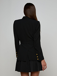 A woman with long brown hair, seen from the back, models the Aven Wrap Blazer—a tailored black wrap-front blazer with gold buttons—paired with a matching pleated skirt against a light background, embodying modern suiting.