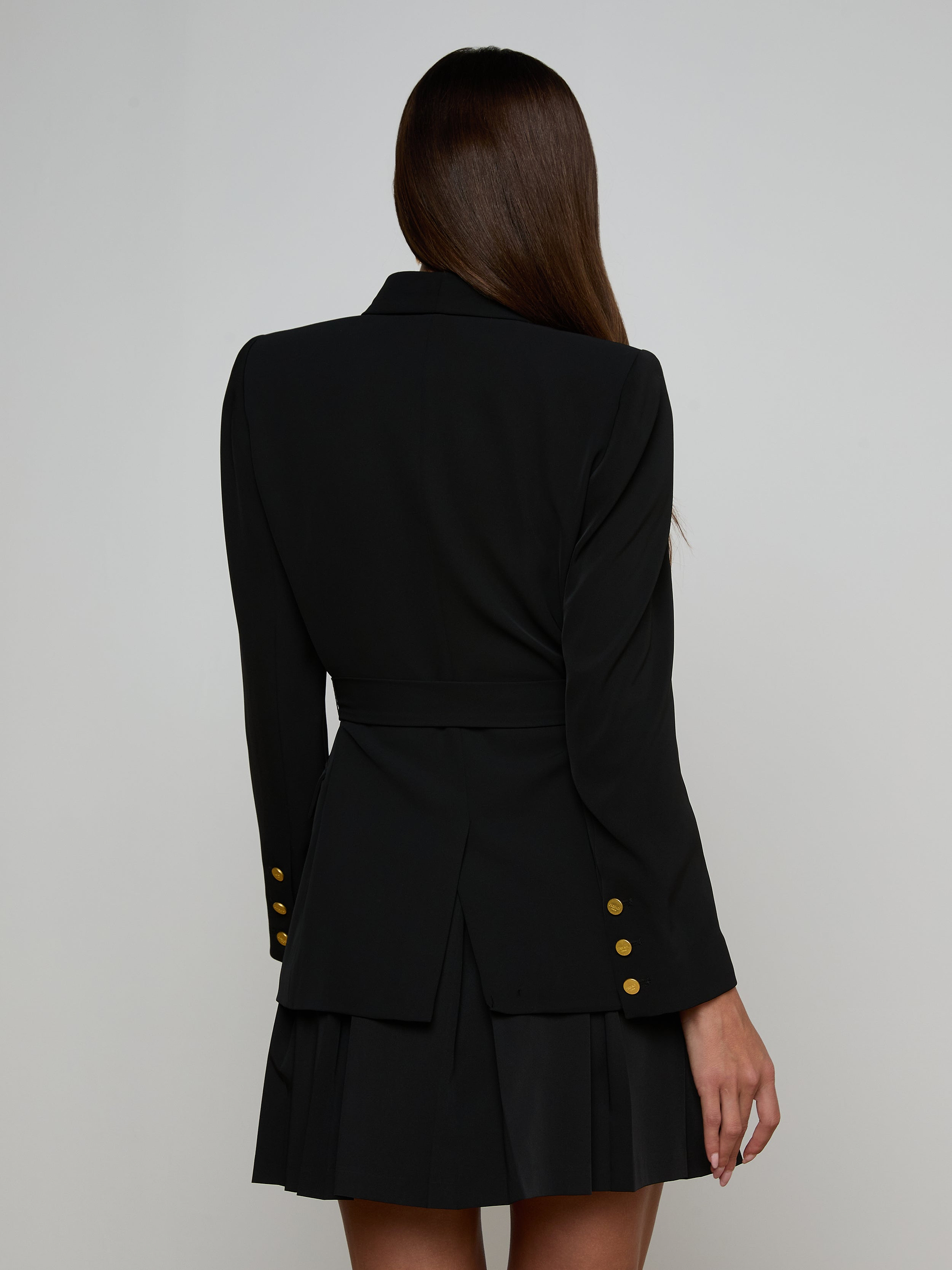 A woman with long brown hair, seen from the back, models the Aven Wrap Blazer—a tailored black wrap-front blazer with gold buttons—paired with a matching pleated skirt against a light background, embodying modern suiting.