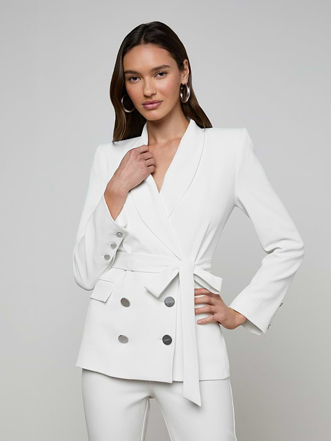A woman poses confidently against a plain light background, wearing the Aven Wrap Blazer—an elegant white wrap-front blazer crafted from modern suiting fabric with silver buttons—paired with matching white pants.