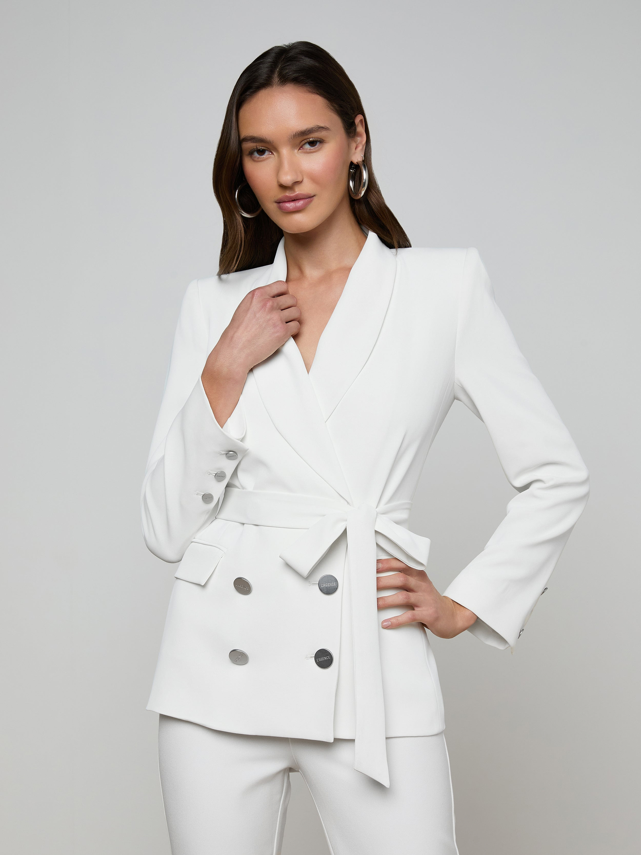 A woman poses confidently against a plain light background, wearing the Aven Wrap Blazer—an elegant white wrap-front blazer crafted from modern suiting fabric with silver buttons—paired with matching white pants.