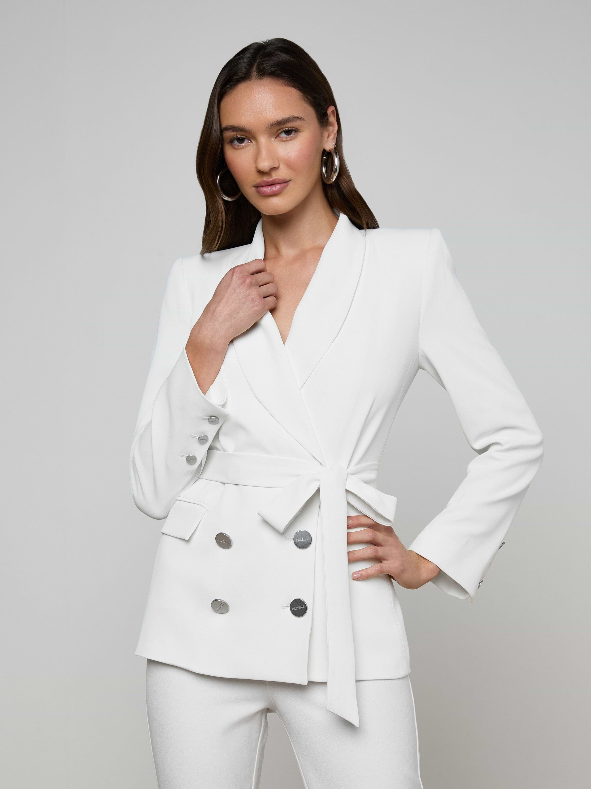 A woman poses confidently against a plain light background, wearing the Aven Wrap Blazer—an elegant white wrap-front blazer crafted from modern suiting fabric with silver buttons—paired with matching white pants.