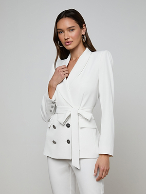A woman with long brown hair wears the Aven Wrap Blazer—a white belted, wrap-front blazer with silver buttons—and matching pants, standing against a light gray background and smiling as she holds her lapel.