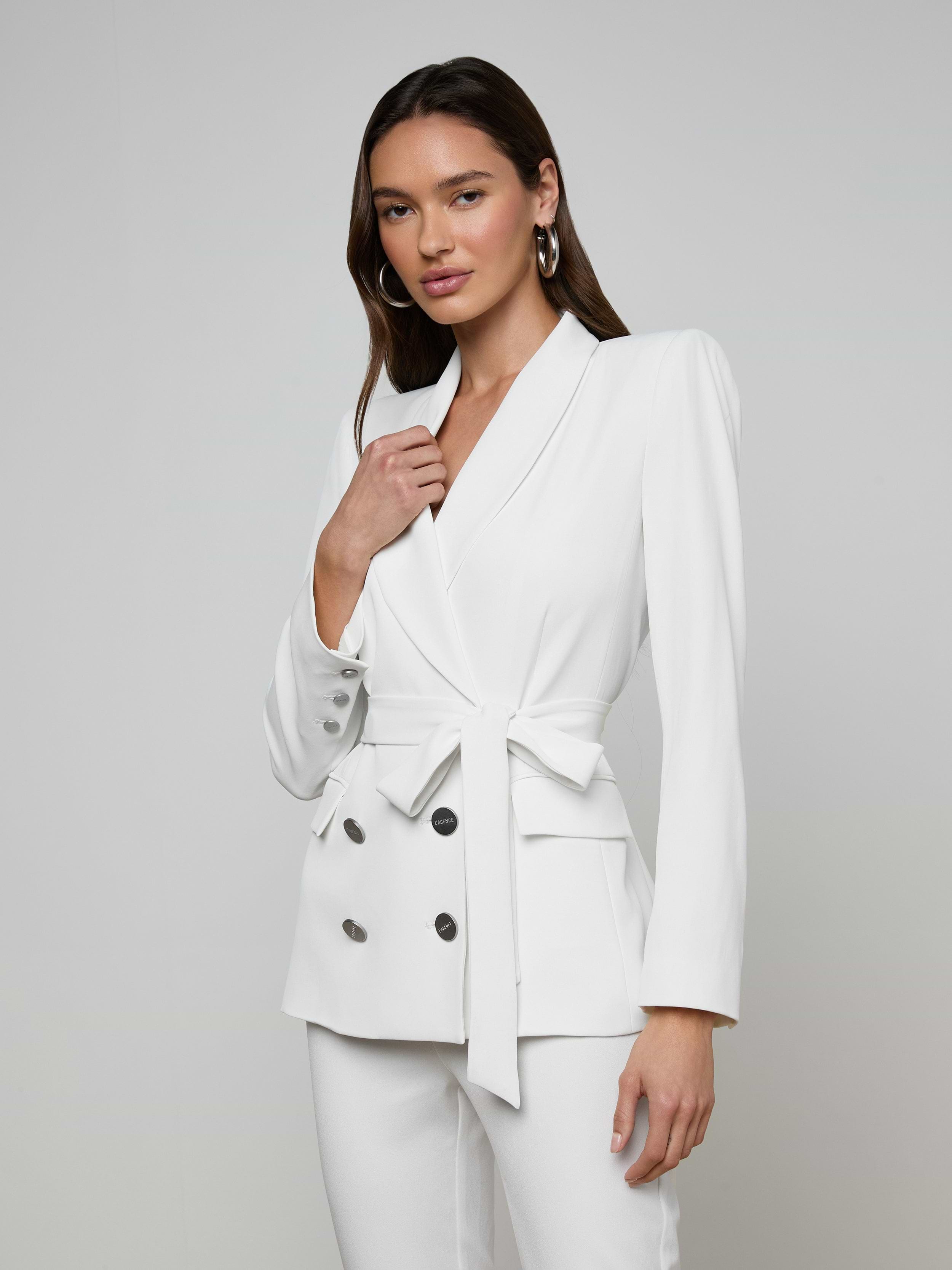 A woman wears the Aven Wrap Blazer—a white tailored pantsuit crafted from modern suiting fabric, featuring a belted waist and double-breasted buttons—standing against a plain light background.