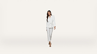 A woman wearing a white suit with a belted blazer and pants stands against a plain white background, looking forward and smiling slightly.