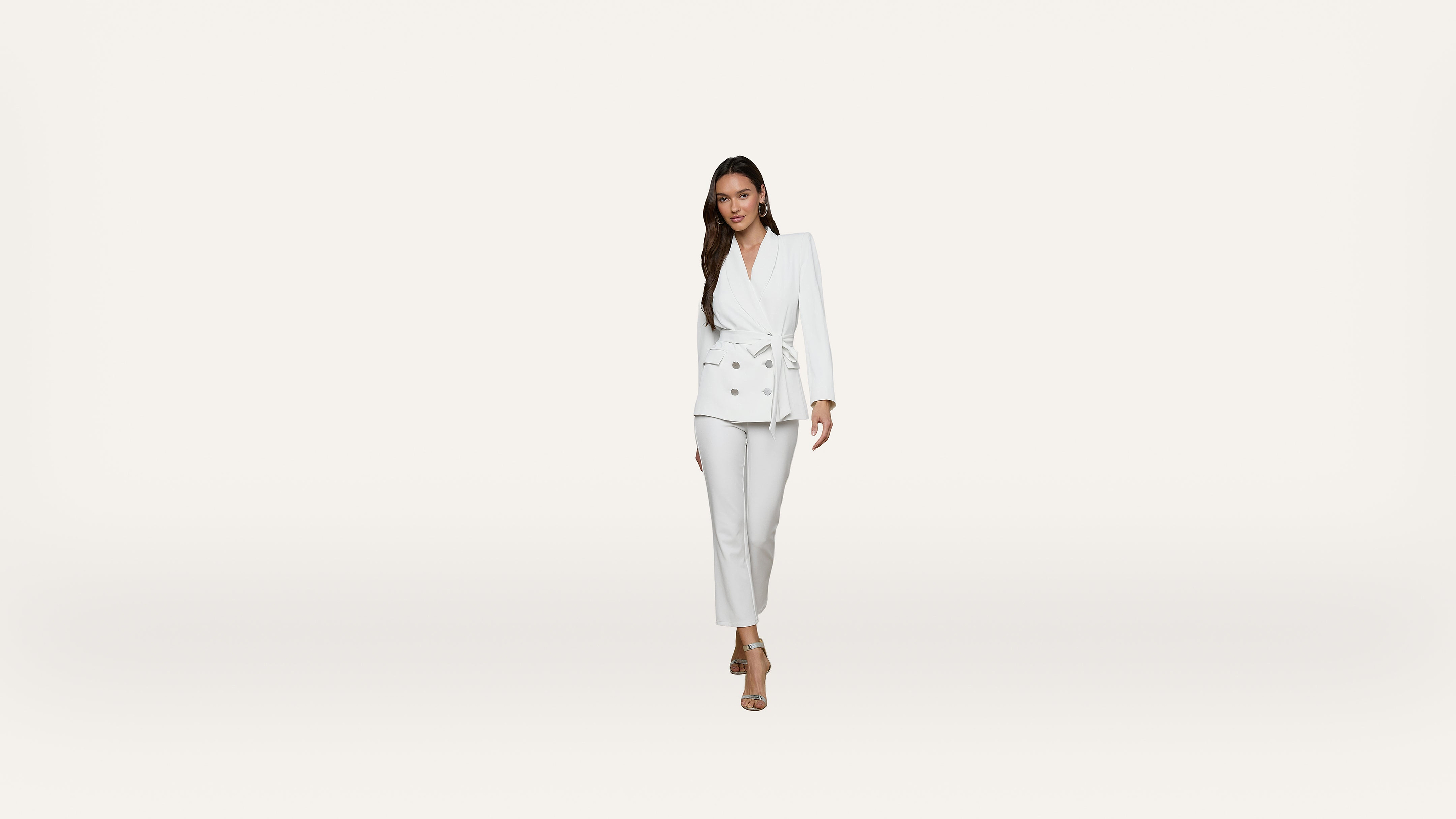 A woman wearing a white suit with a belted blazer and pants stands against a plain white background, looking forward and smiling slightly.