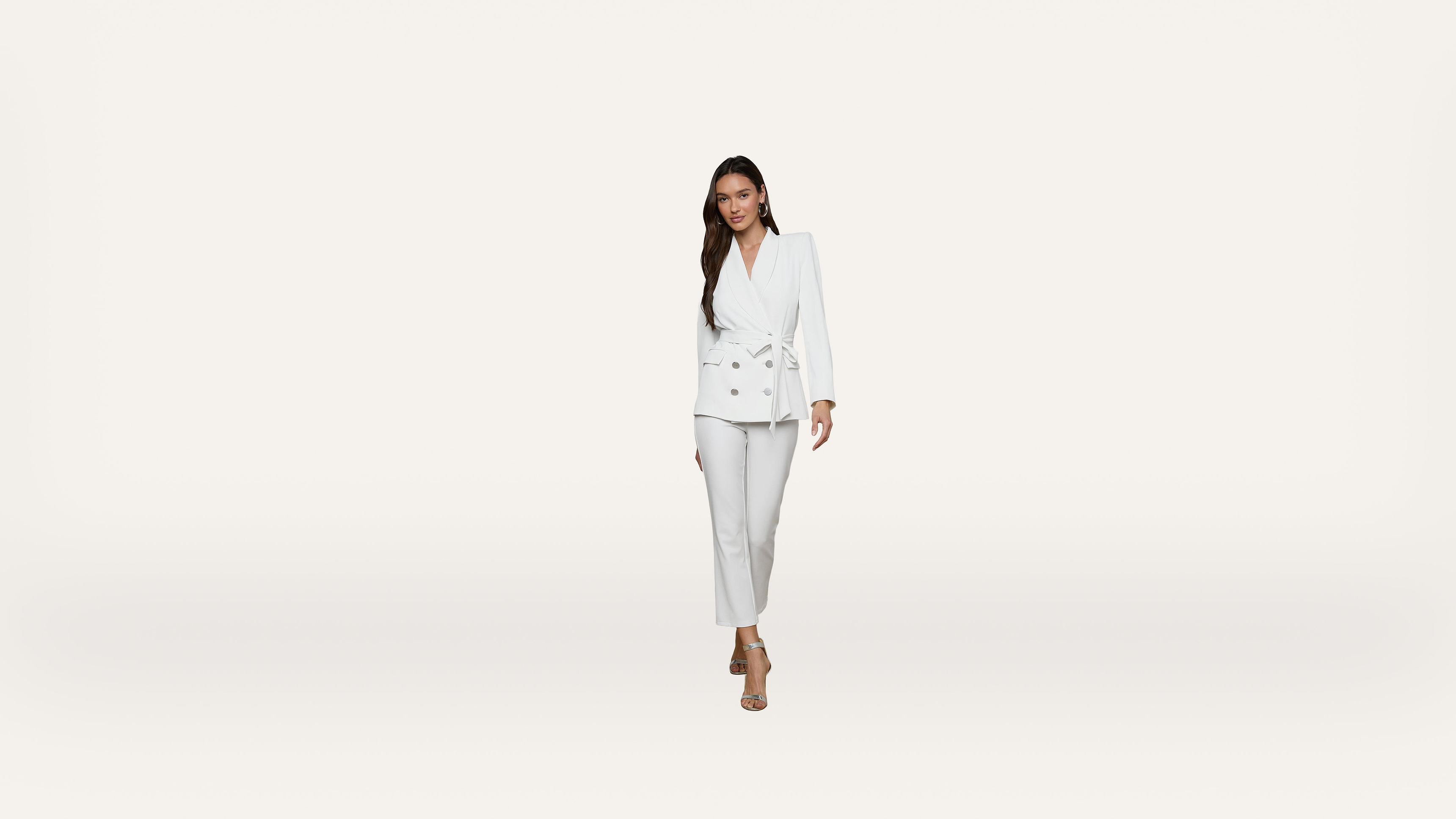 A woman wearing a white suit with a belted blazer and pants stands against a plain white background, looking forward and smiling slightly.