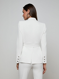 A woman stands with her back turned, dressed in the Aven Wrap Blazer—a fitted white wrap-front blazer with a belt and matching white pants, crafted from modern suiting fabric. The jacket showcases button details on the sleeves. She is set against a plain, light-colored background.