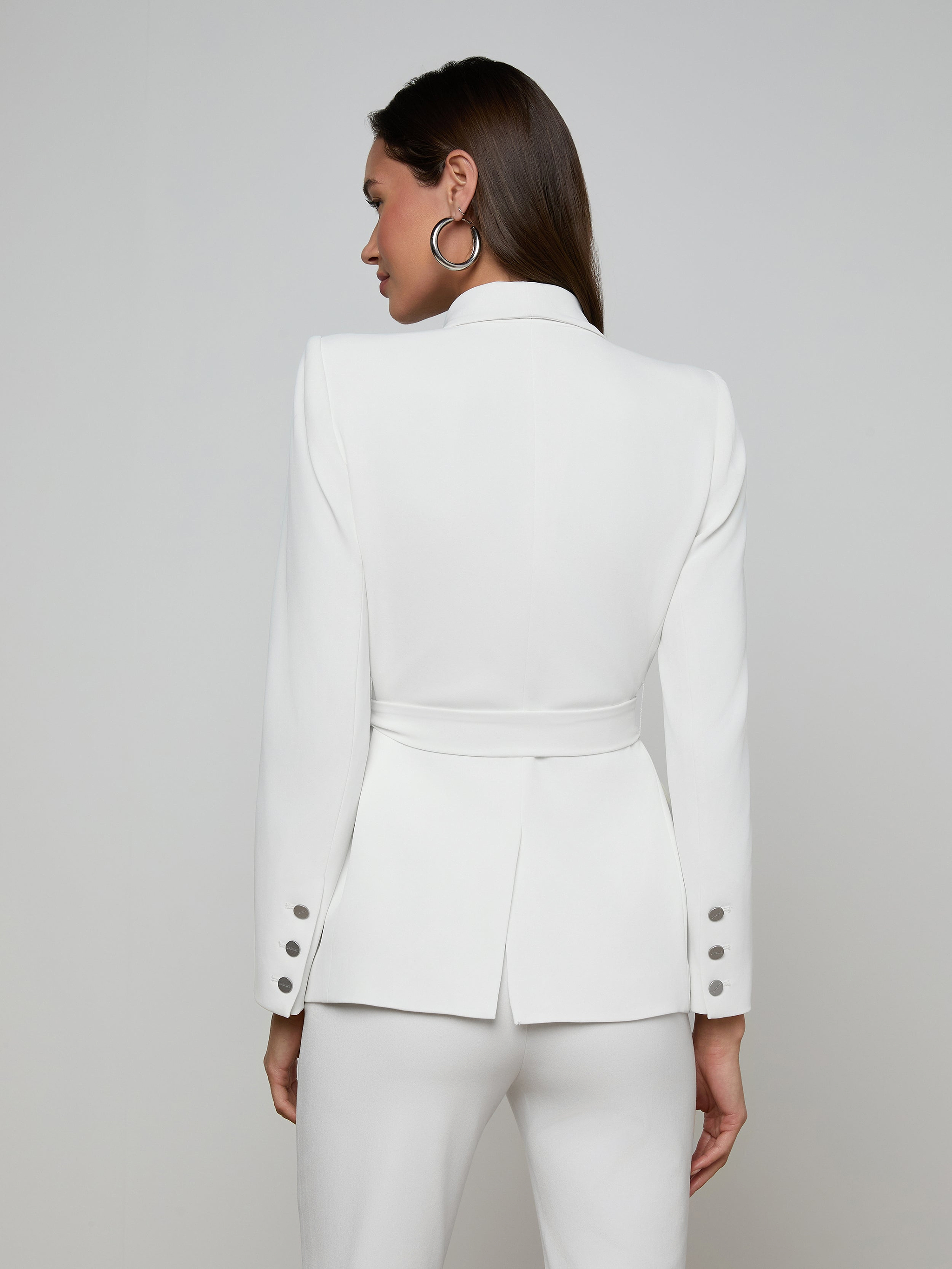 A woman stands with her back turned, dressed in the Aven Wrap Blazer—a fitted white wrap-front blazer with a belt and matching white pants, crafted from modern suiting fabric. The jacket showcases button details on the sleeves. She is set against a plain, light-colored background.