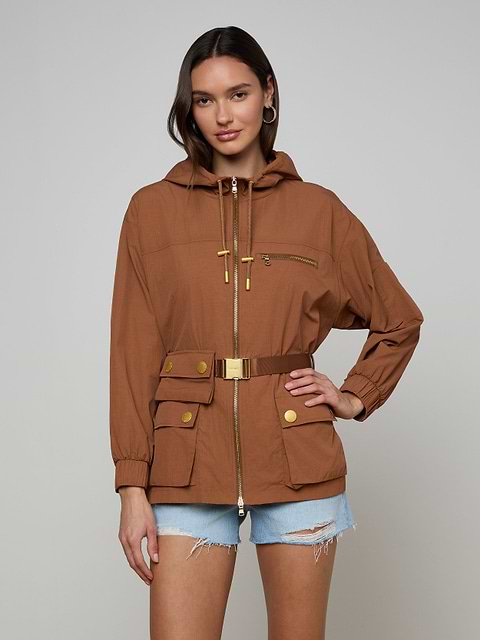 Wearing the Calina Windbreaker—a brown, utility-chic hooded jacket with gold zippers and buttons and a belted waist—she pairs it with light blue distressed denim shorts, posing against a plain light background.