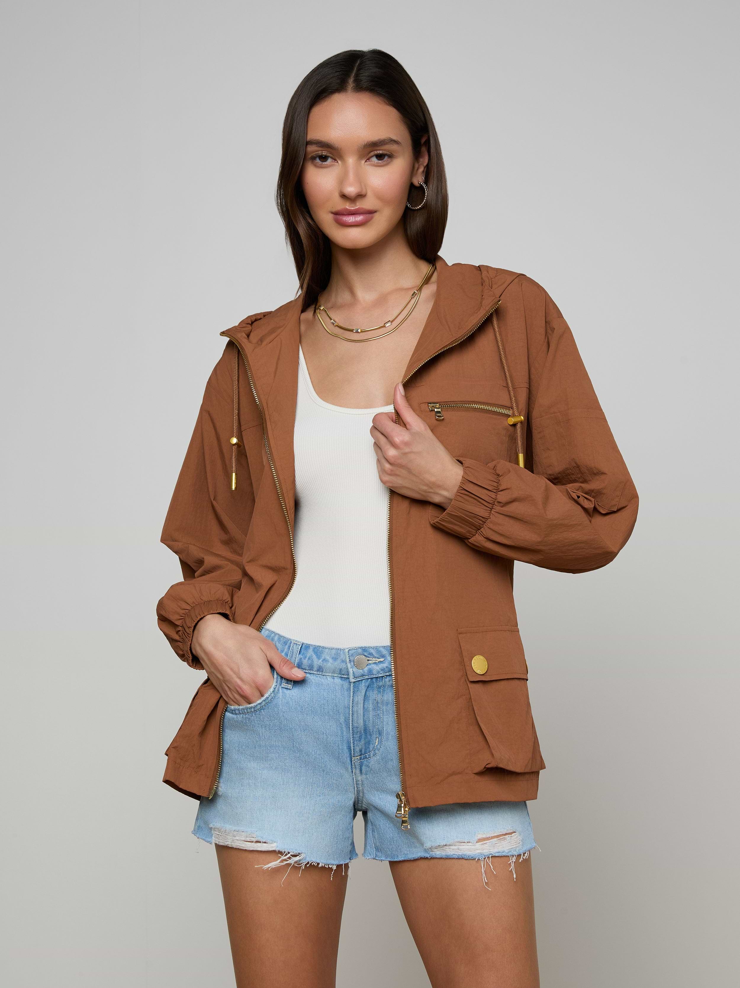 A woman stands against a plain light background with one hand in her pocket, sporting the Calina Windbreaker in brown over a white top and light blue denim shorts for a chic utility-inspired look.
