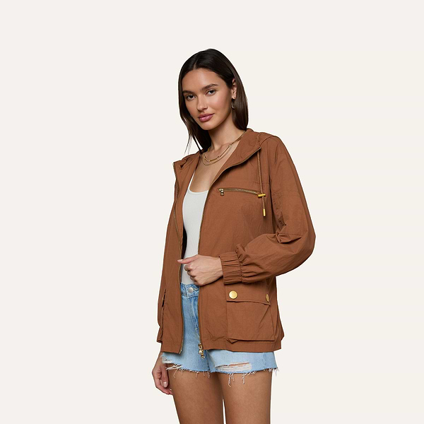 Wearing the brown Calina Windbreaker layered over a white top and denim shorts, a woman stands against a plain light background, looking at the camera with a slight smile.