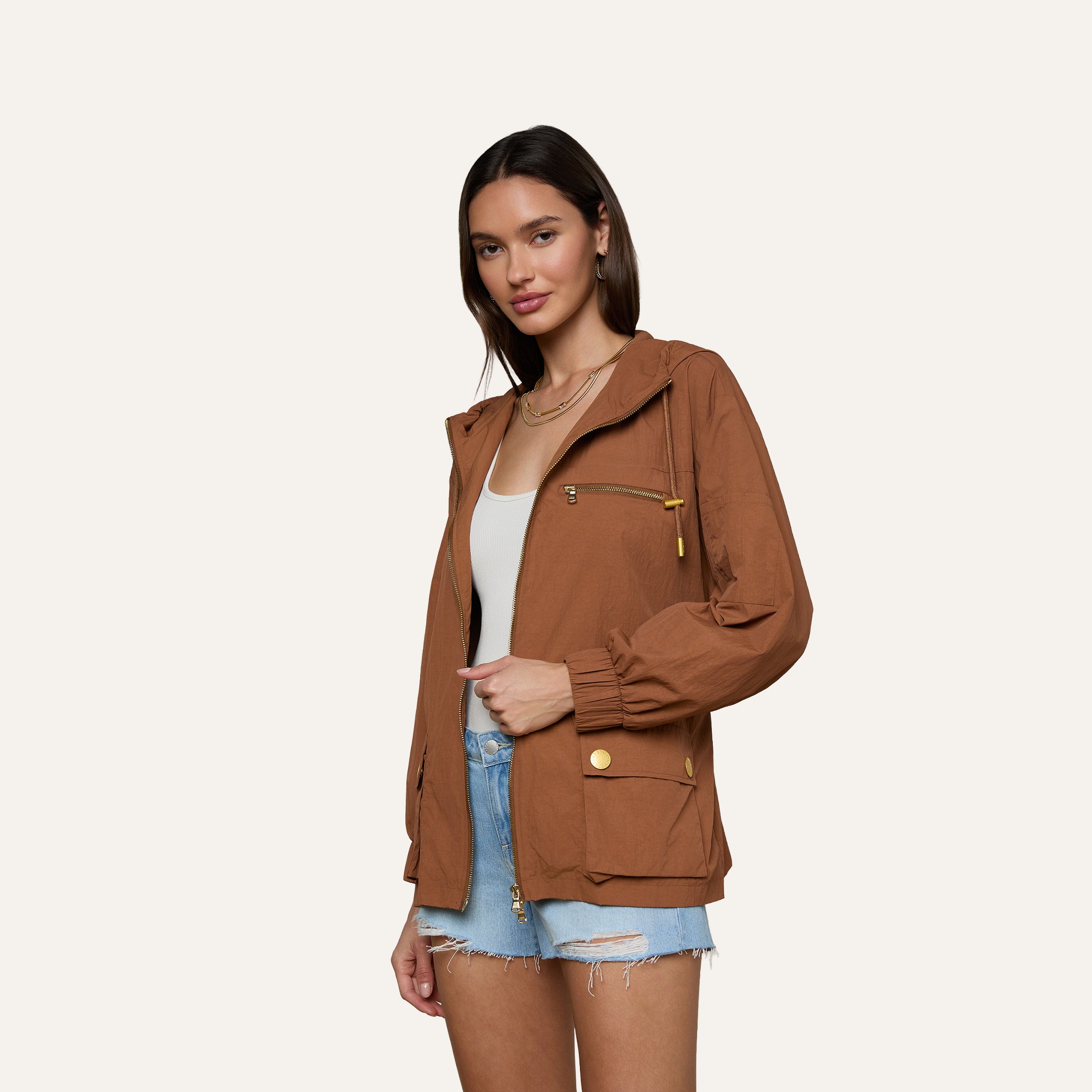 Wearing the brown Calina Windbreaker layered over a white top and denim shorts, a woman stands against a plain light background, looking at the camera with a slight smile.