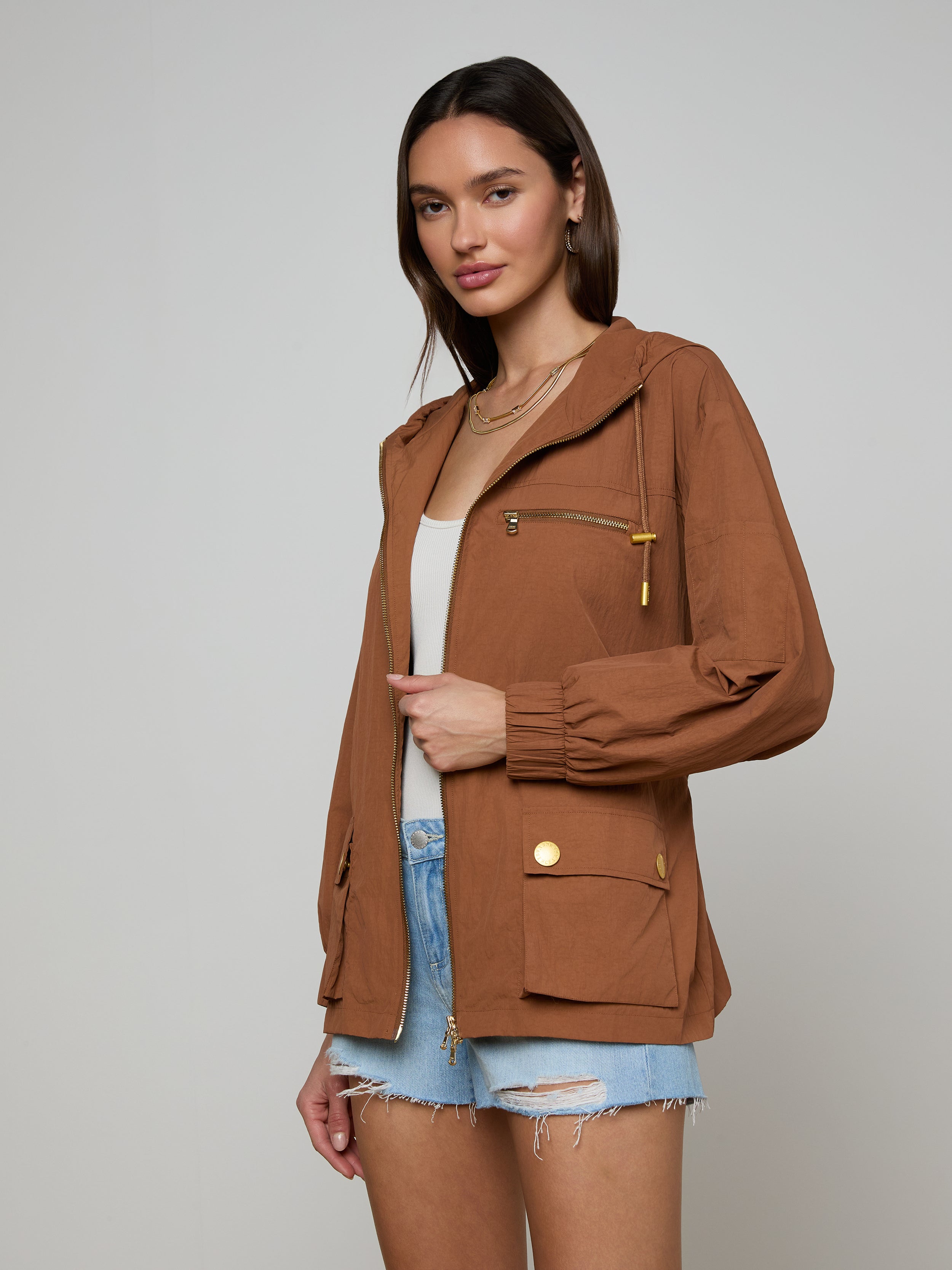 A woman stands against a plain light background, wearing the Calina Windbreaker in brown over a white top and distressed denim shorts.