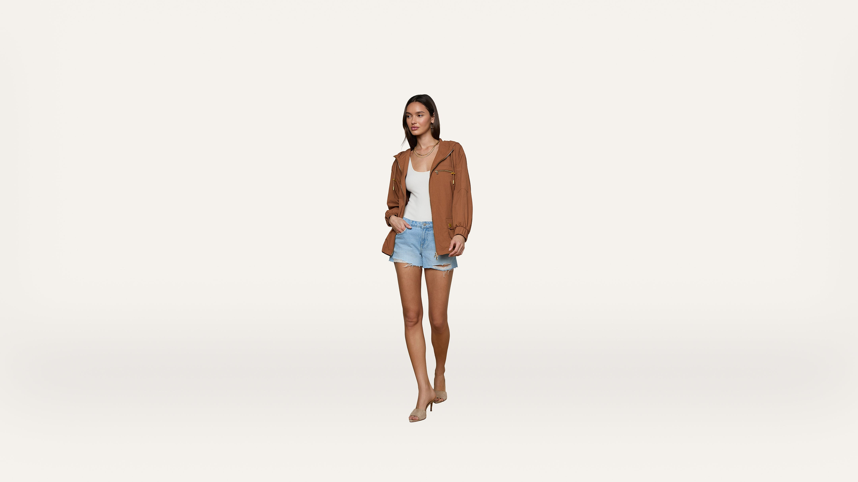 A woman walks forward wearing a brown jacket, white top, light blue shorts, and nude heels against a plain white background.