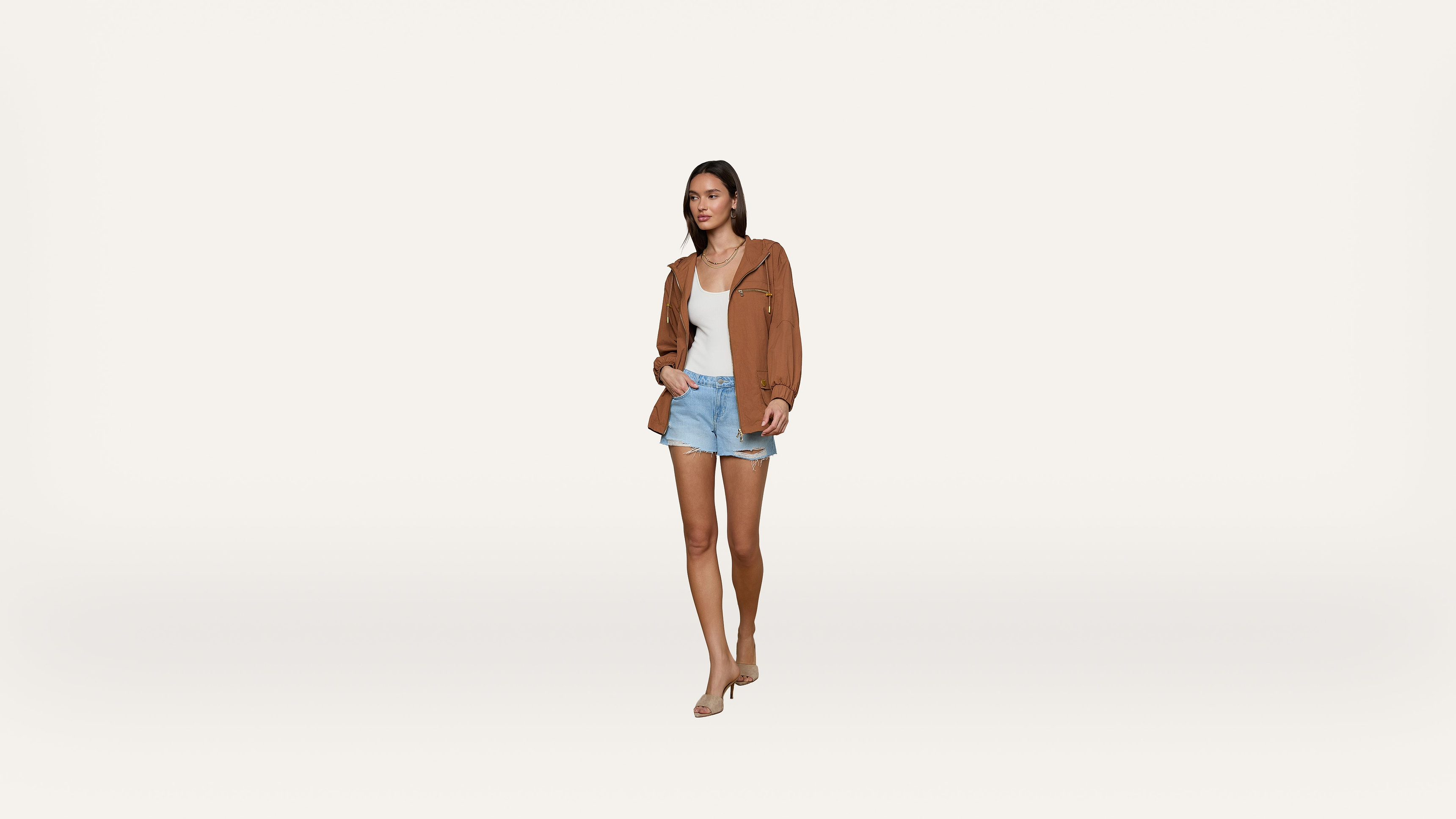 A woman walks forward wearing a brown jacket, white top, light blue shorts, and nude heels against a plain white background.