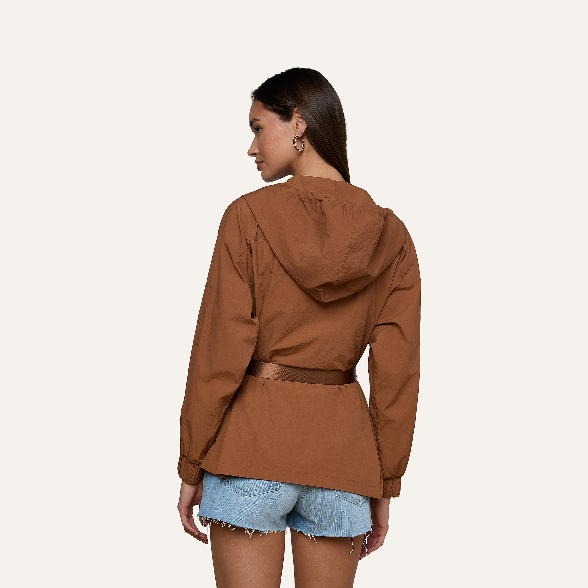 A woman with long brown hair stands with her back to the camera, wearing the Calina Windbreaker in brown—complete with a belt—and light blue denim shorts. She poses against a plain, light background, embodying a utility-chic style.