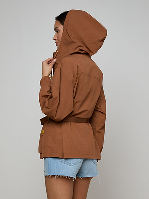 A person in a Calina Windbreaker with a hood and light denim shorts stands sideways against a plain light background, looking ahead while touching the hood.