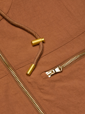 Close-up of the Calina Windbreaker, a brown utility jacket with a gold zipper, small zippered pocket, and brown drawstring with gold metal tips. This water-repellent design combines style and functionality.