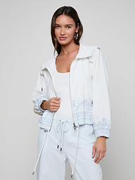 Woman wearing the Miro Hooded Windbreaker in white with light blue patterns, paired with white drawstring pants and a white top, standing against a plain light background.