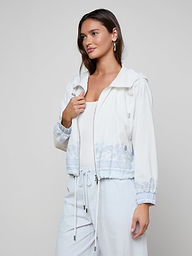 A woman stands against a plain light background, looking slightly to the side, wearing the Miro Hooded Windbreaker in white with light blue patterns over a white top and white pants.