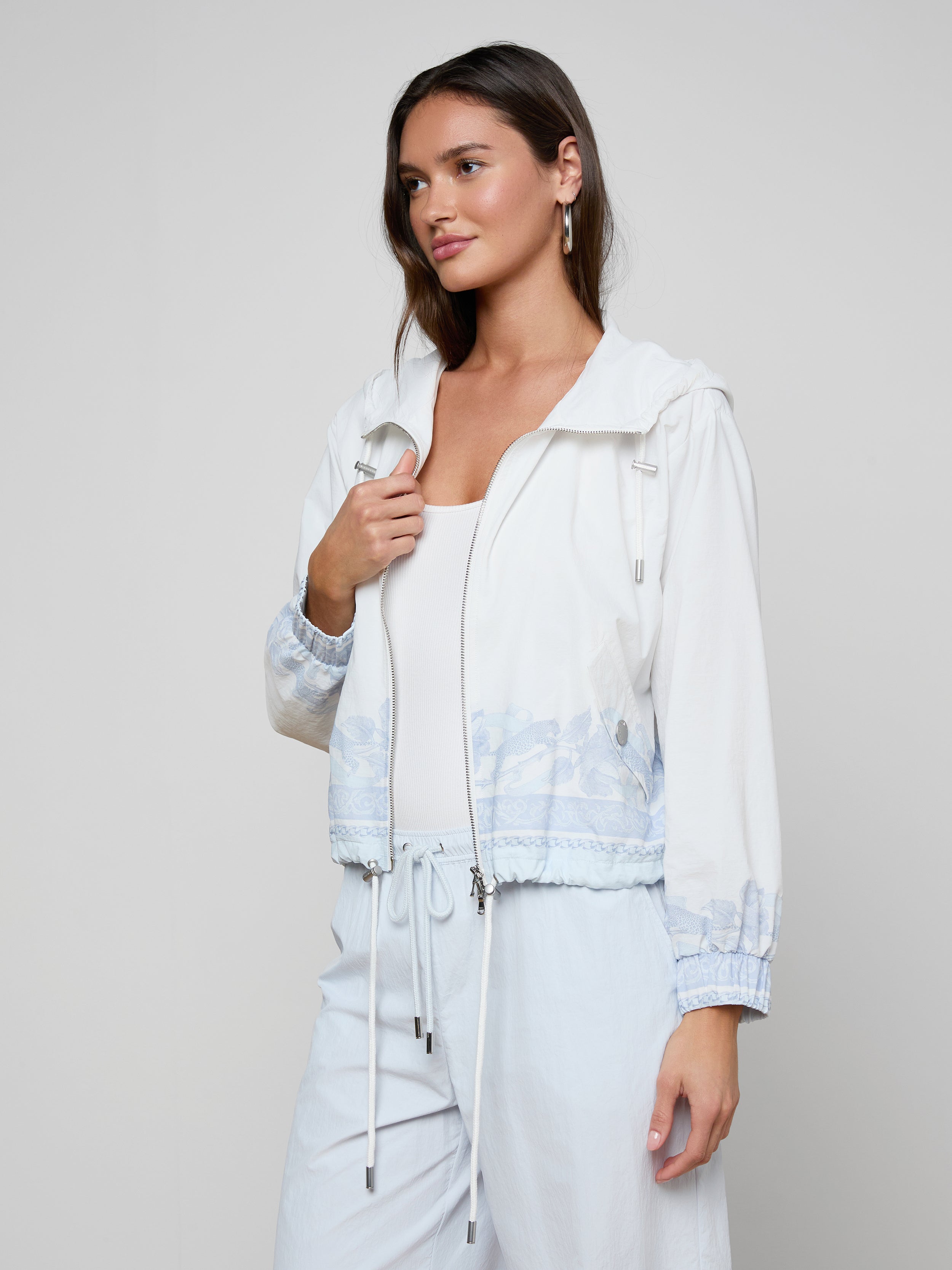A woman stands against a plain light background, looking slightly to the side, wearing the Miro Hooded Windbreaker in white with light blue patterns over a white top and white pants.