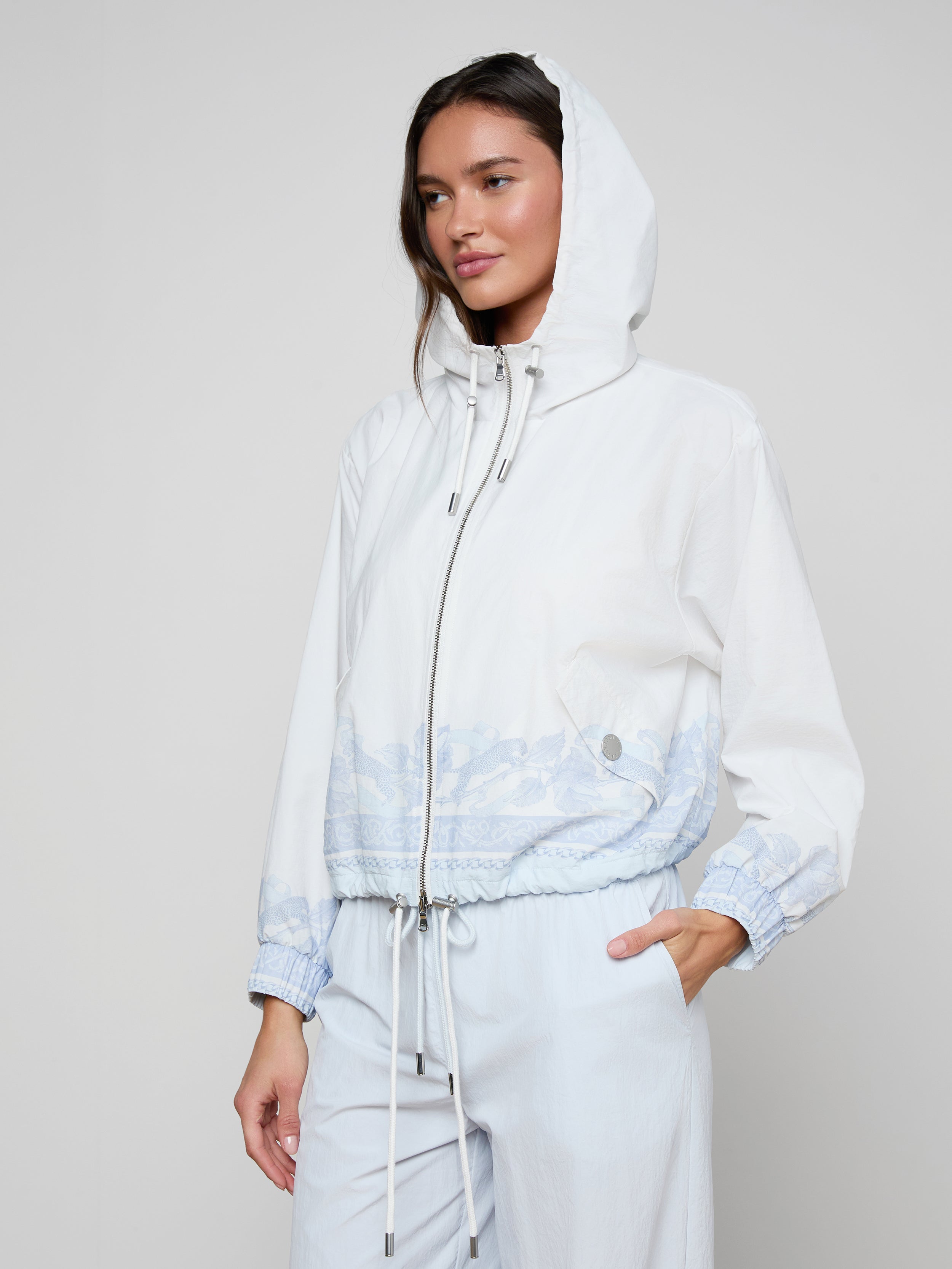 A woman is wearing the Miro Hooded Windbreaker in white with light blue patterns near the waist and cuffs, paired with light blue pants. She has one hand in her pocket and is standing against a plain background.
