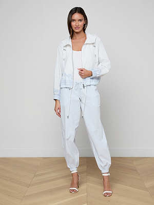 A woman stands indoors on a light wood floor, wearing a white Miro Hooded Windbreaker over a white top and high-waisted loose white pants with elastic cuffs, paired with white high-heeled sandals. The background is plain.