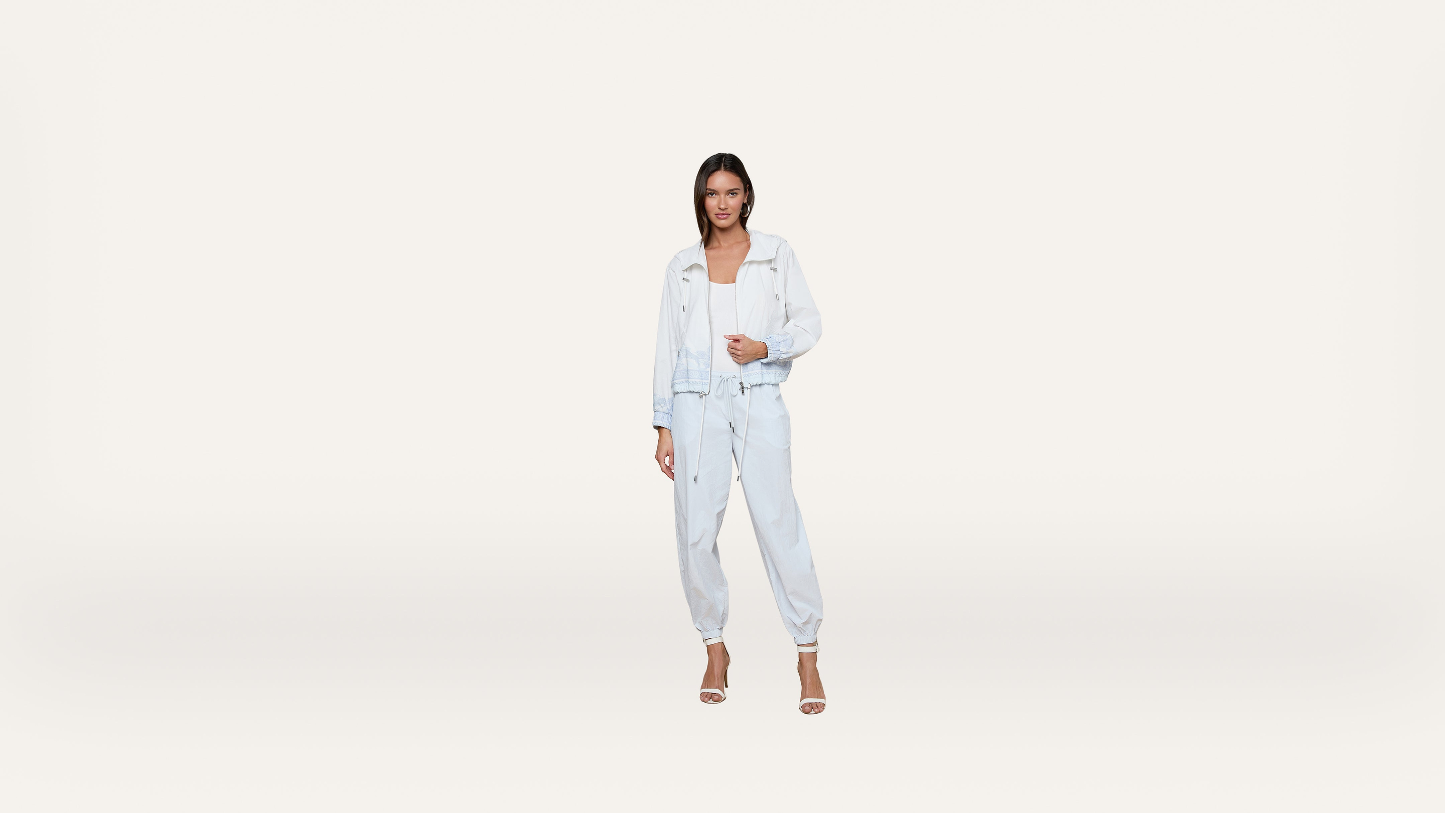 A woman wearing a light blue denim jacket and jogger pants with a white top and heels stands against a plain white background.