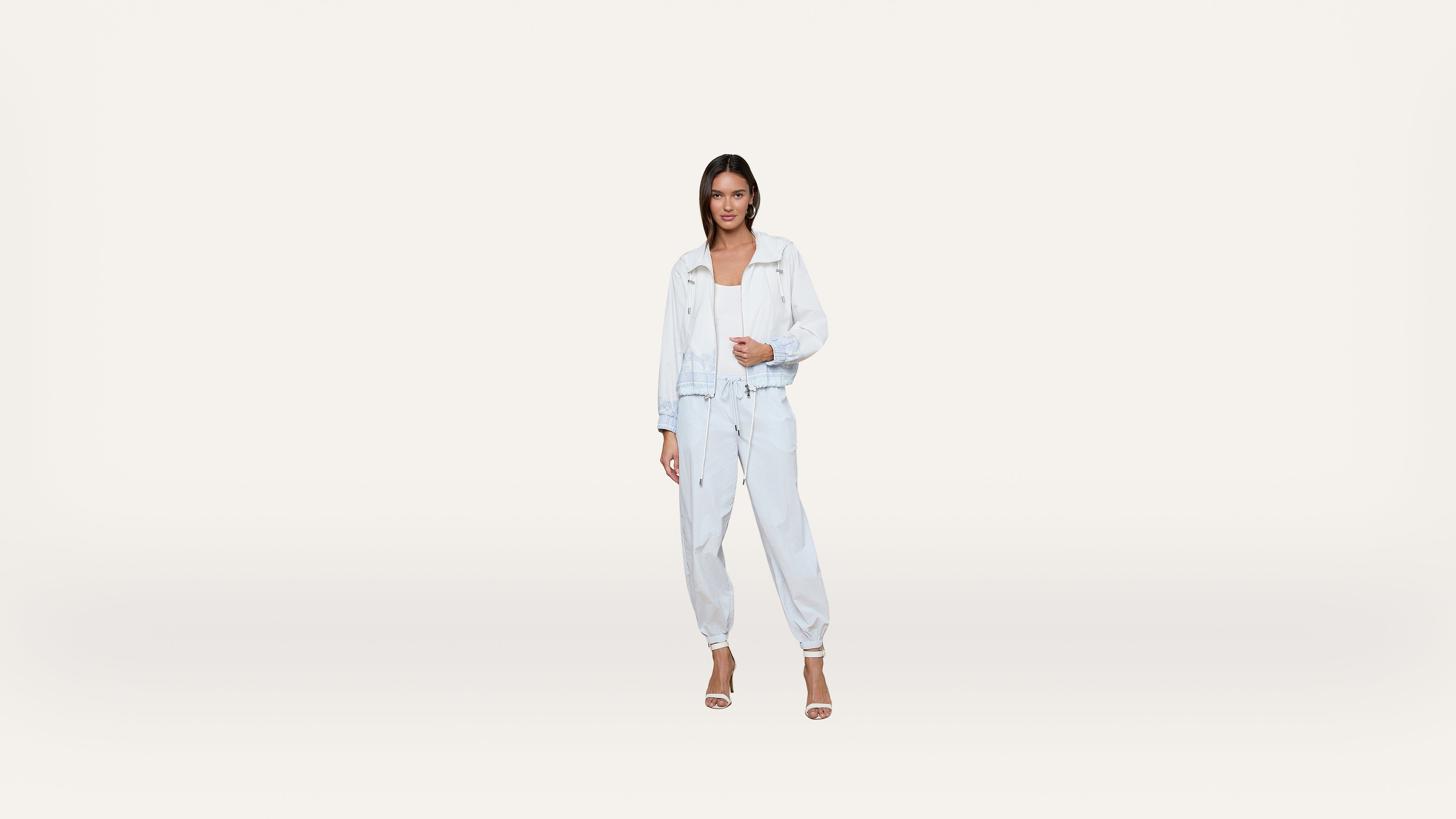 A woman wearing a light blue denim jacket and jogger pants with a white top and heels stands against a plain white background.
