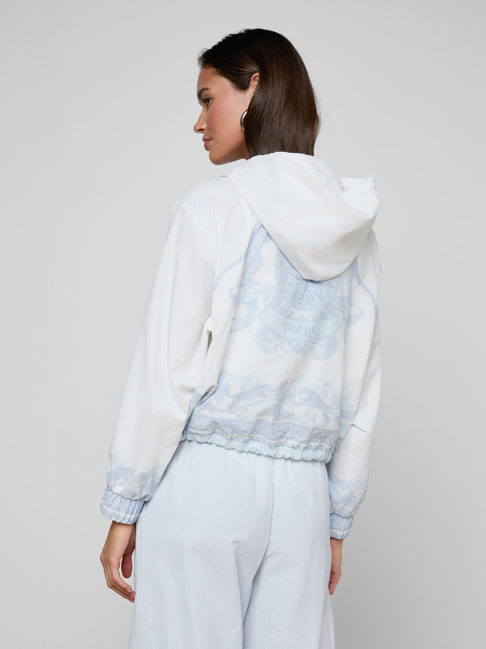 A woman stands facing away against an off-white background, wearing the Miro Hooded Windbreaker in white with pale blue designs and light blue pants.