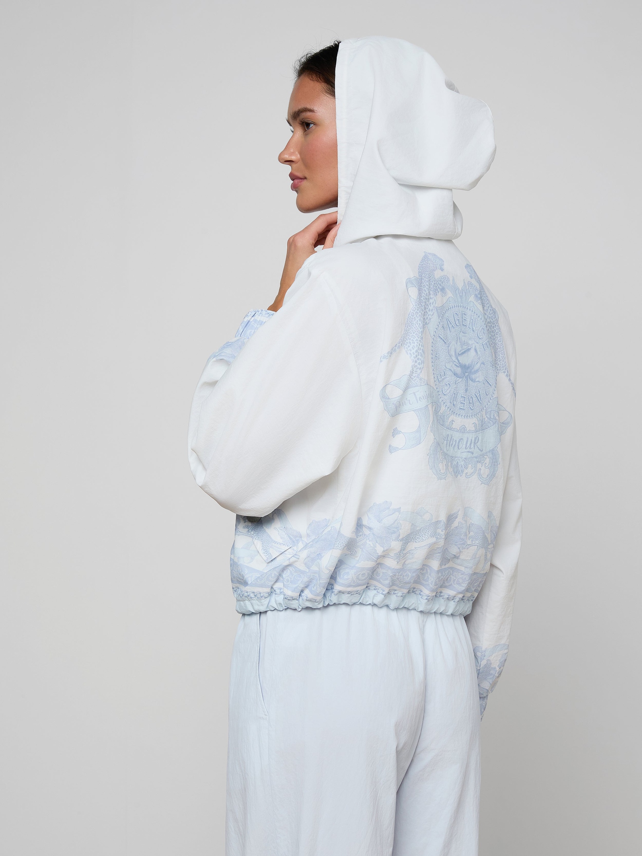 Wearing the Miro Hooded Windbreaker in white with a light blue design on the back and matching light blue pants, a person stands sideways against a plain background.