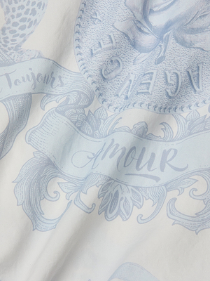 A close-up of the Miro Hooded Windbreaker’s white fabric, adorned with light blue floral patterns, swirls, a statement logo, and French words like Toujours and Amour in elegant cursive.