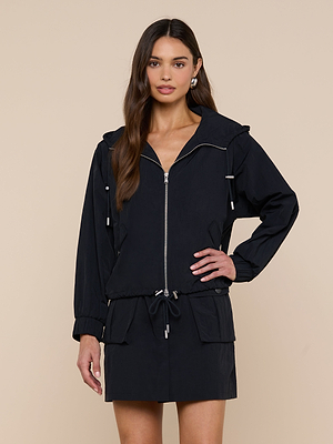 Una mujer de pelo largo y castaño luce el modelo Miro Hooded Windbreaker, una chaqueta negra con cremallera y cintura con cordón, combinada con una falda corta a juego con bolsillos delanteros, posando sobre un fondo beige con una mano en la cadera.