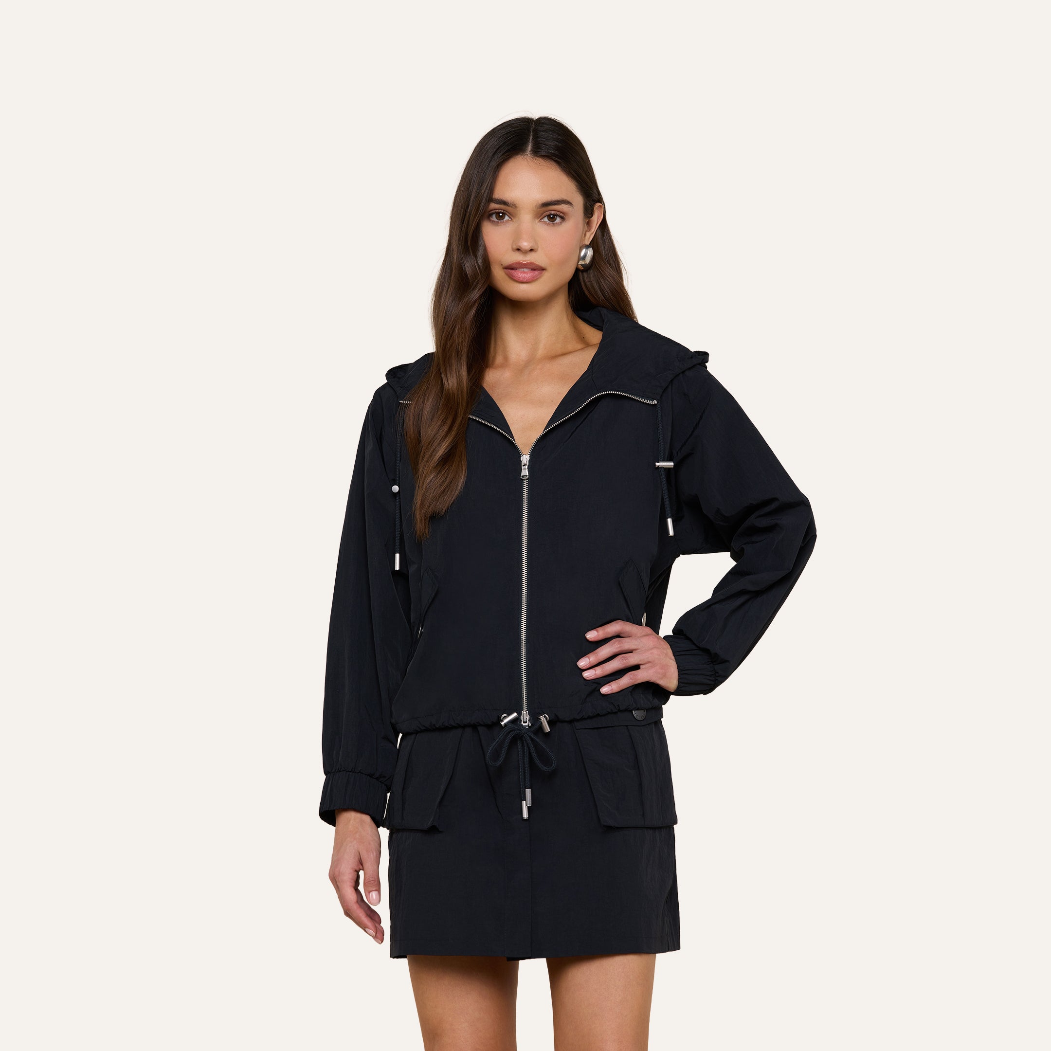 A woman with long brown hair wears the Miro Hooded Windbreaker, featuring a zip closure, drawstring waist, and front pockets, paired with a matching black skirt while standing against a plain light background.