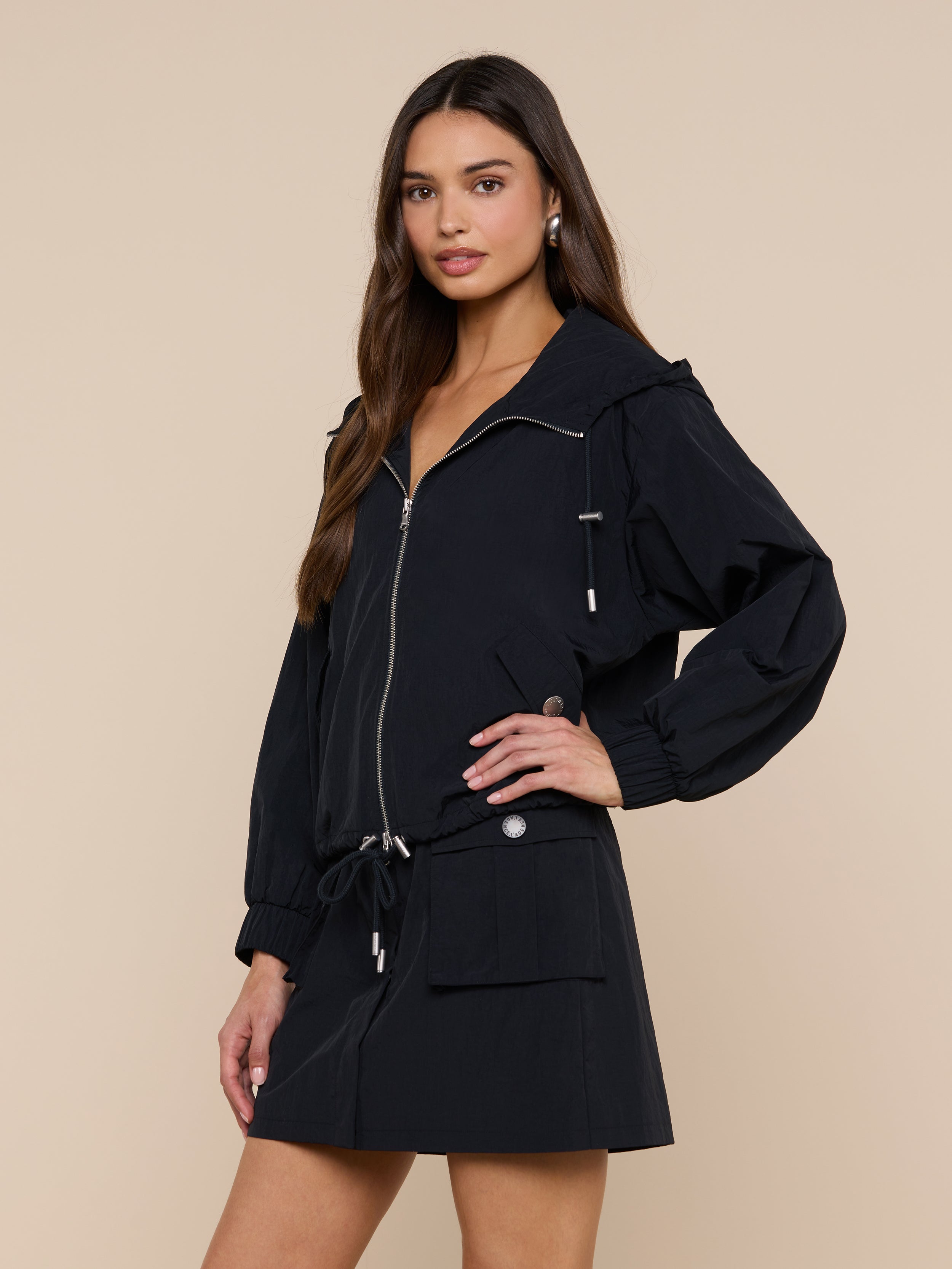 A woman wearing the Miro Hooded Windbreaker with zip closure and a matching black skirt stands against a plain light background, looking at the camera.