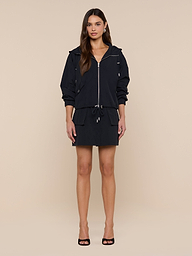 A woman stands facing forward wearing the Miro Hooded Windbreaker with zip closure and a matching drawstring waist skirt, paired with black open-toe heels against a plain white background.