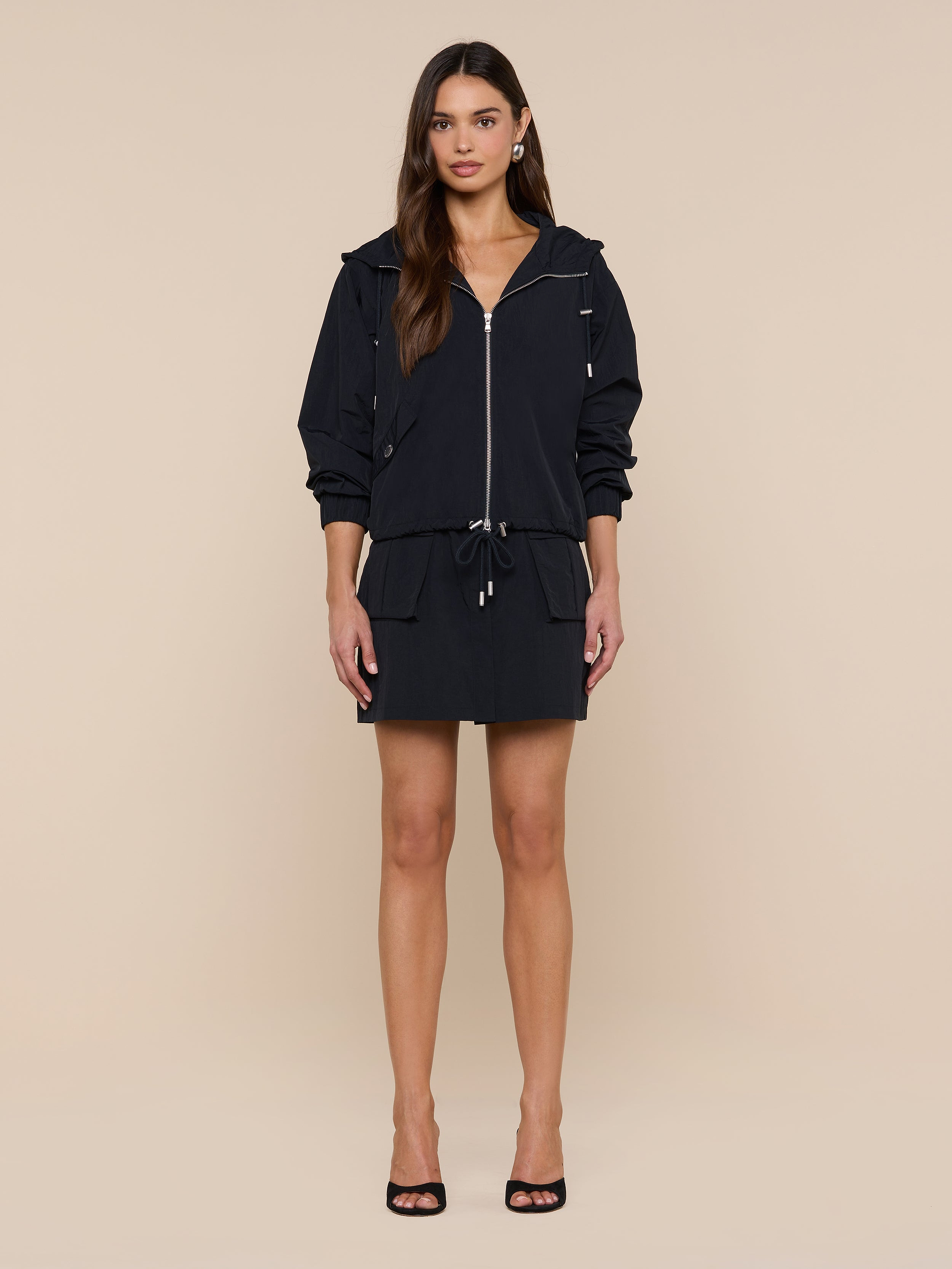 A woman stands facing forward wearing the Miro Hooded Windbreaker with zip closure and a matching drawstring waist skirt, paired with black open-toe heels against a plain white background.