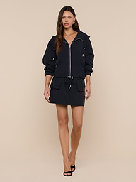 A woman stands against a plain white background, wearing the Miro Hooded Windbreaker—a black zip-up dress with a drawstring waist—paired with black heels. Featuring a sleek zip closure, the look is complemented by her long brown hair and neutral expression.