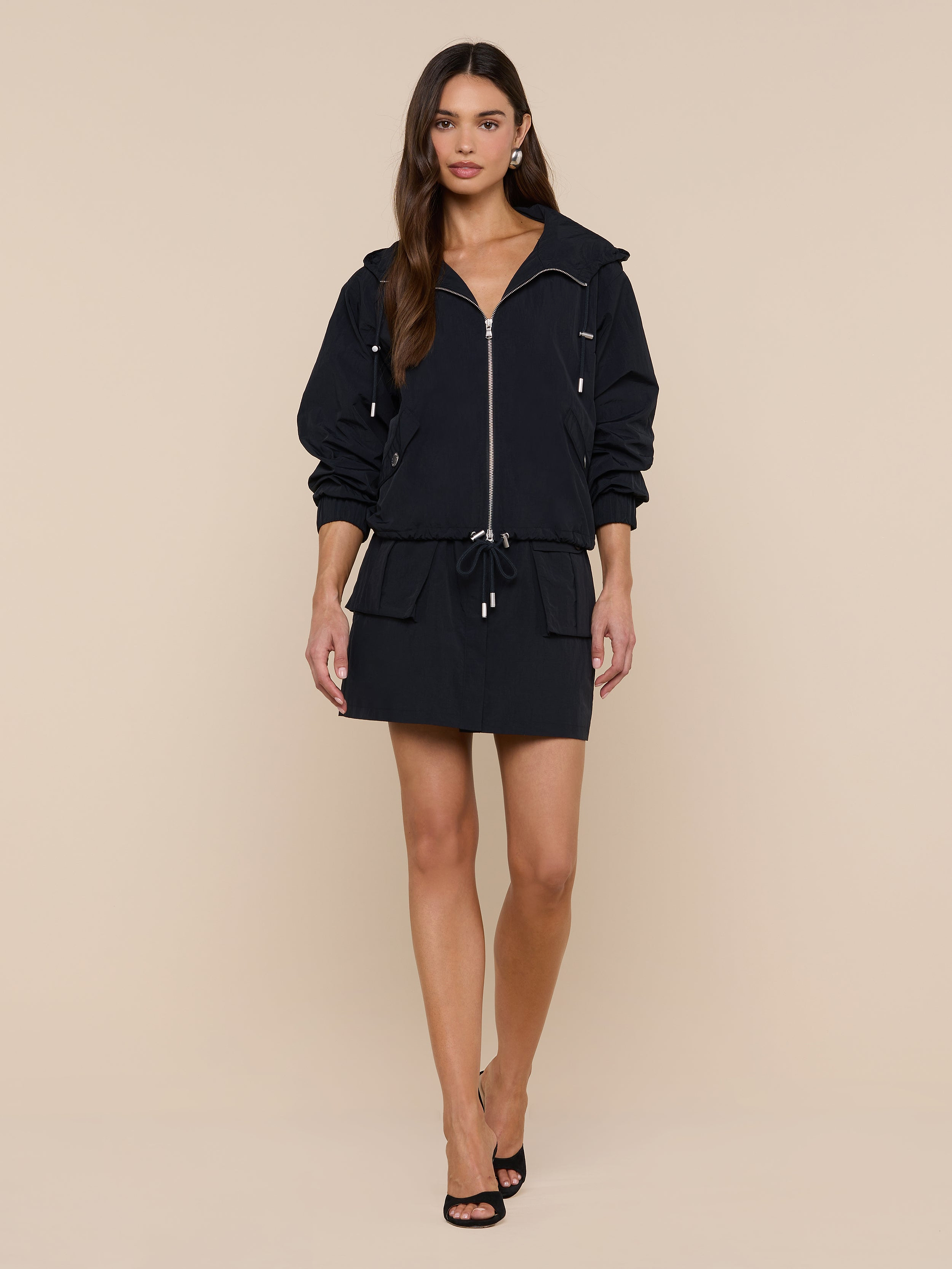 A woman stands against a plain white background, wearing the Miro Hooded Windbreaker—a black zip-up dress with a drawstring waist—paired with black heels. Featuring a sleek zip closure, the look is complemented by her long brown hair and neutral expression.