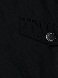 A close-up of a black fabric pocket with a flap and a metallic button engraved with text, featured on the Miro Hooded Windbreaker. The button is attached near the edge of the water-repellent pocket.