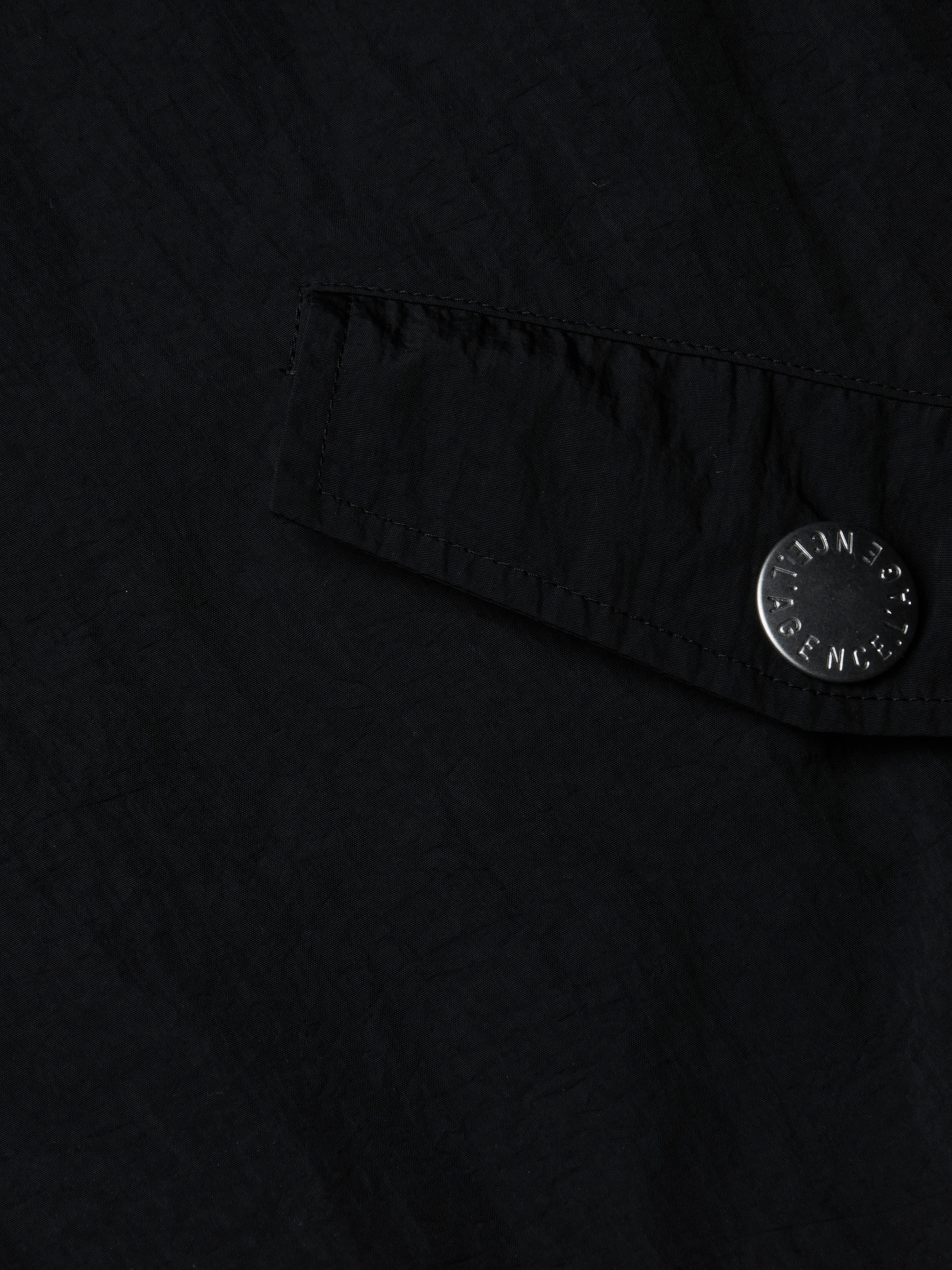 A close-up of a black fabric pocket with a flap and a metallic button engraved with text, featured on the Miro Hooded Windbreaker. The button is attached near the edge of the water-repellent pocket.