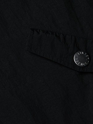 Un primer plano del cortavientos con capucha Miro muestra un tejido negro con solapa y un botón redondo con el grabado STONE ISLAND. El material ligeramente arrugado y repelente al agua resalta el diseño elevado y protector de la chaqueta.