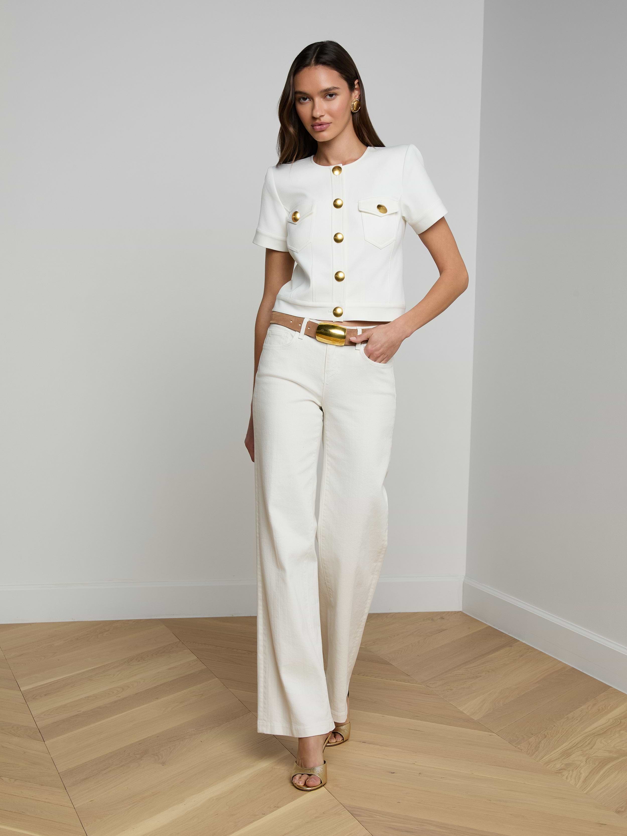 A woman stands against a plain background wearing the Nawa Shirt Jacket—a tailored white short-sleeve button-up top with gold buttons—paired with white wide-leg pants, a white belt with a gold buckle, and beige heels.
