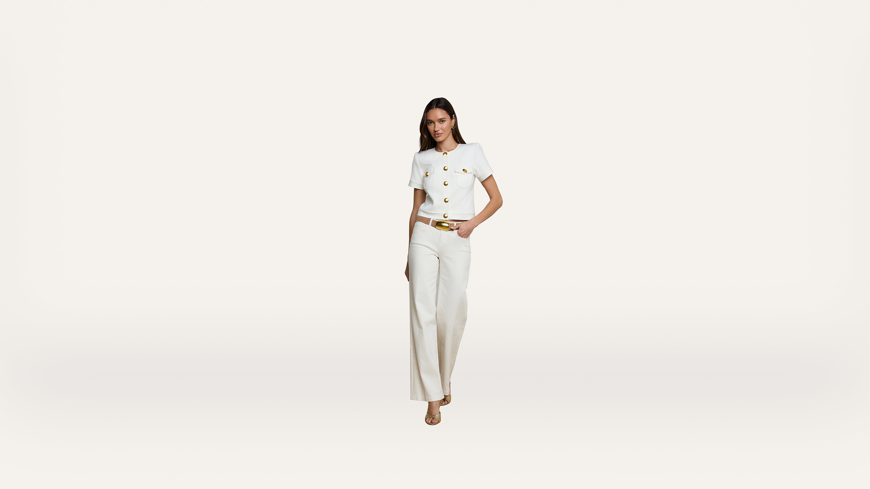 A woman wearing a white short-sleeved top with gold buttons and white high-waisted trousers stands against a plain white background. She has one hand in her pocket and is wearing beige heels.